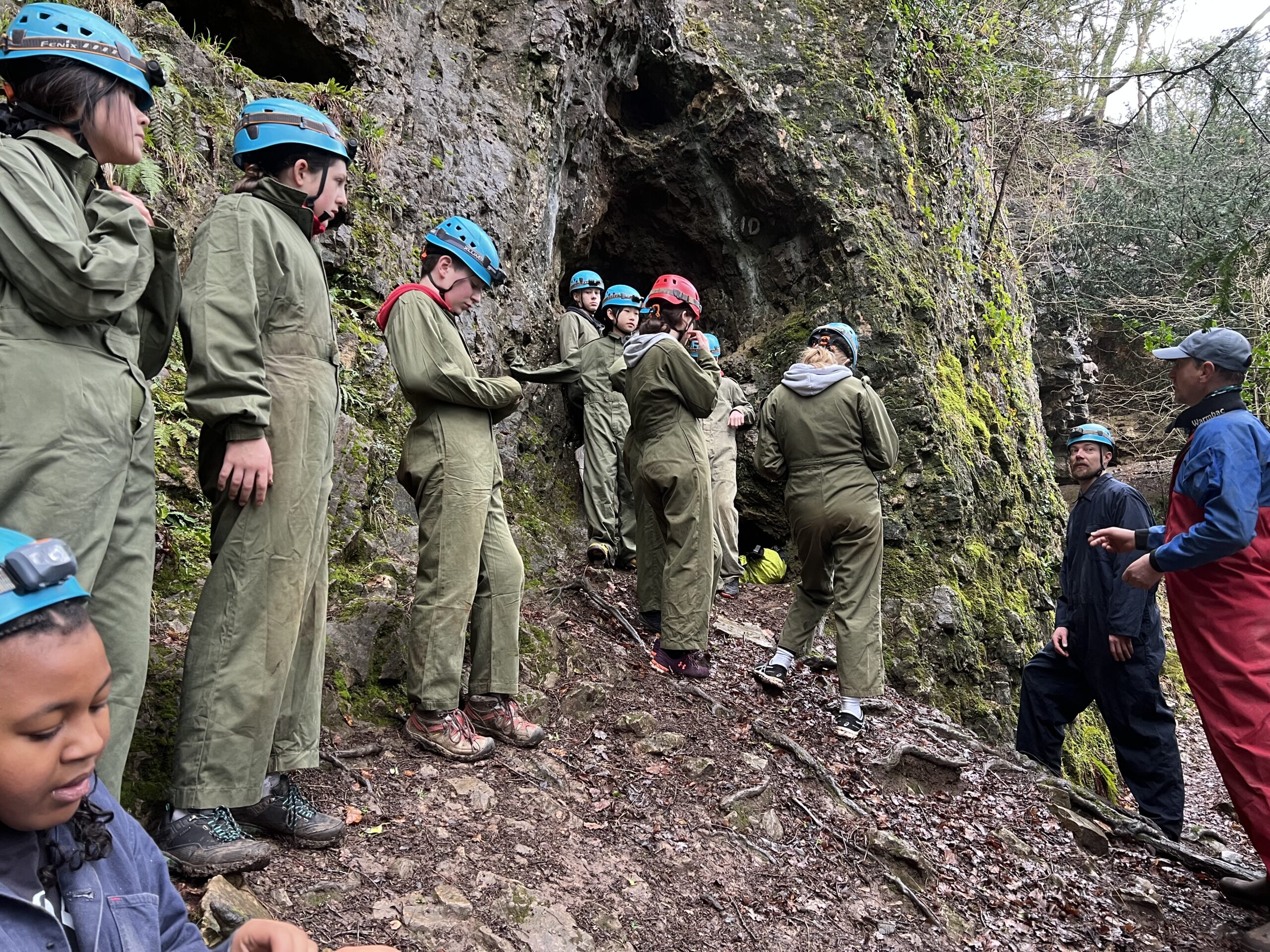 A group of children in caving equipment stood outside a cave entrance.