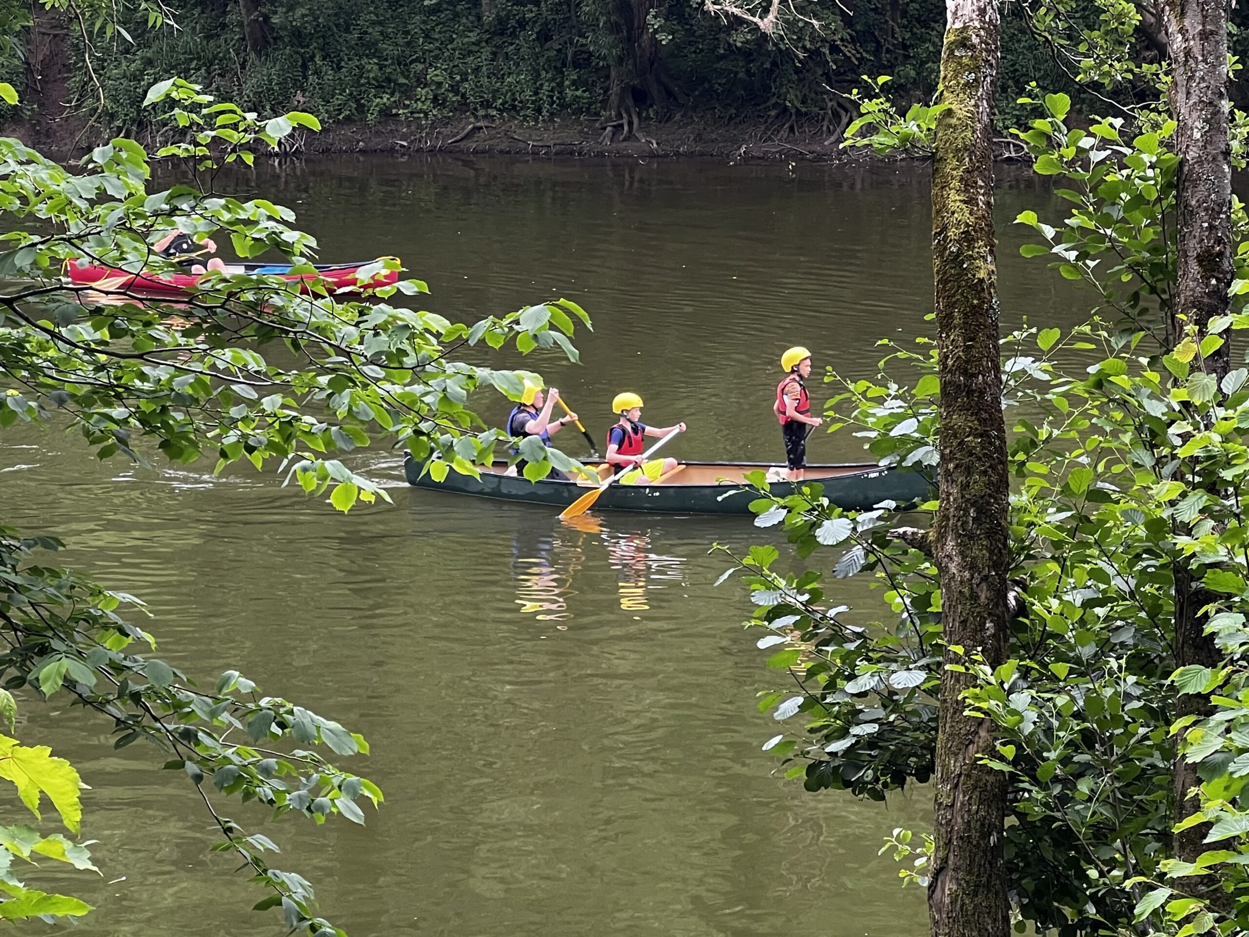 Through the trees a green canoe and red canoe on a river with children aboard.
