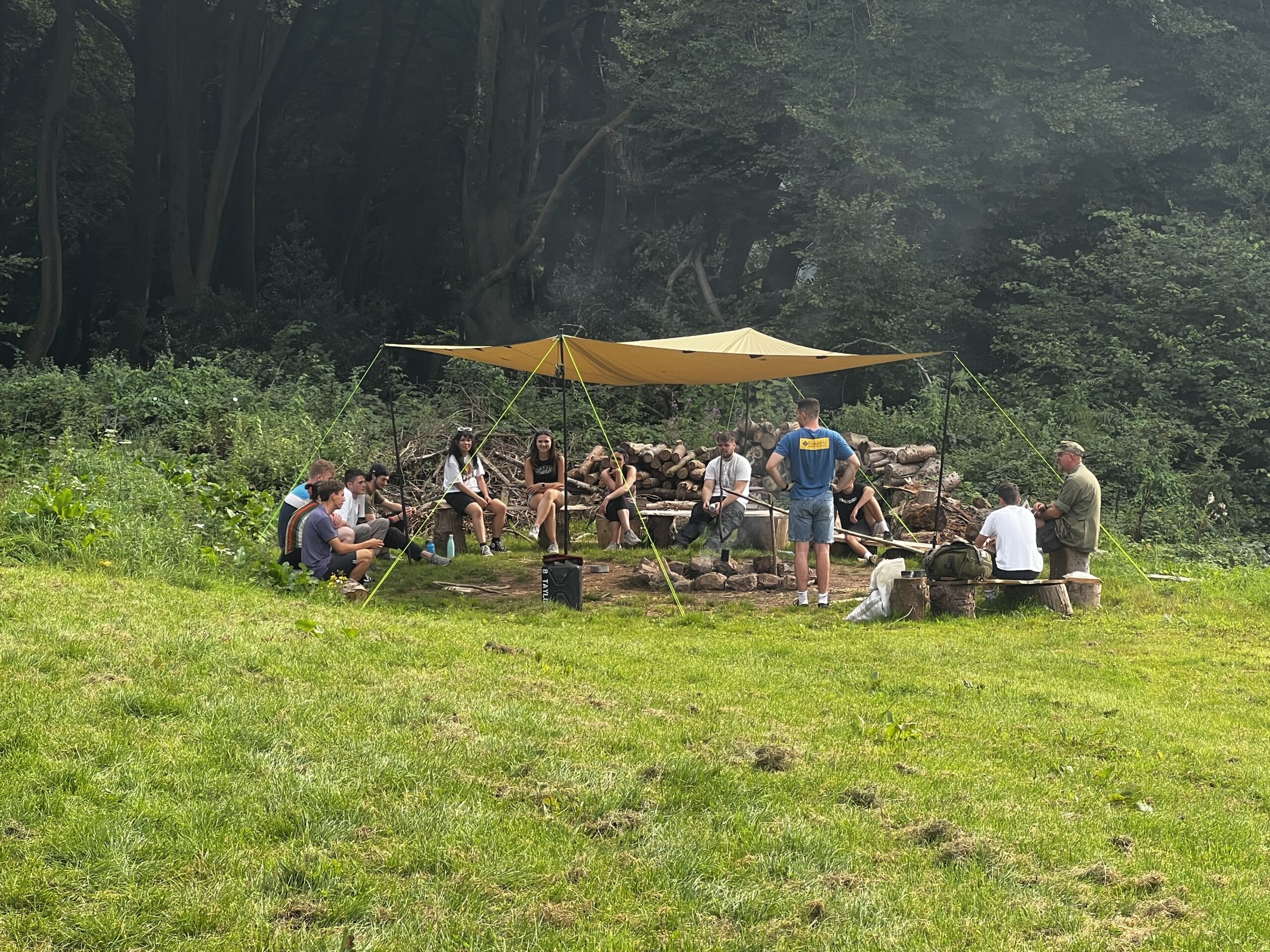 A group of people sat on tree stumps around a fire pit under a shelter.