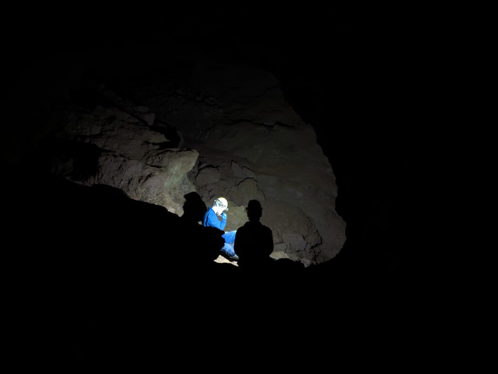 A dark cave with a man in blue overalls and hard hat shining a torch down a tunnel.