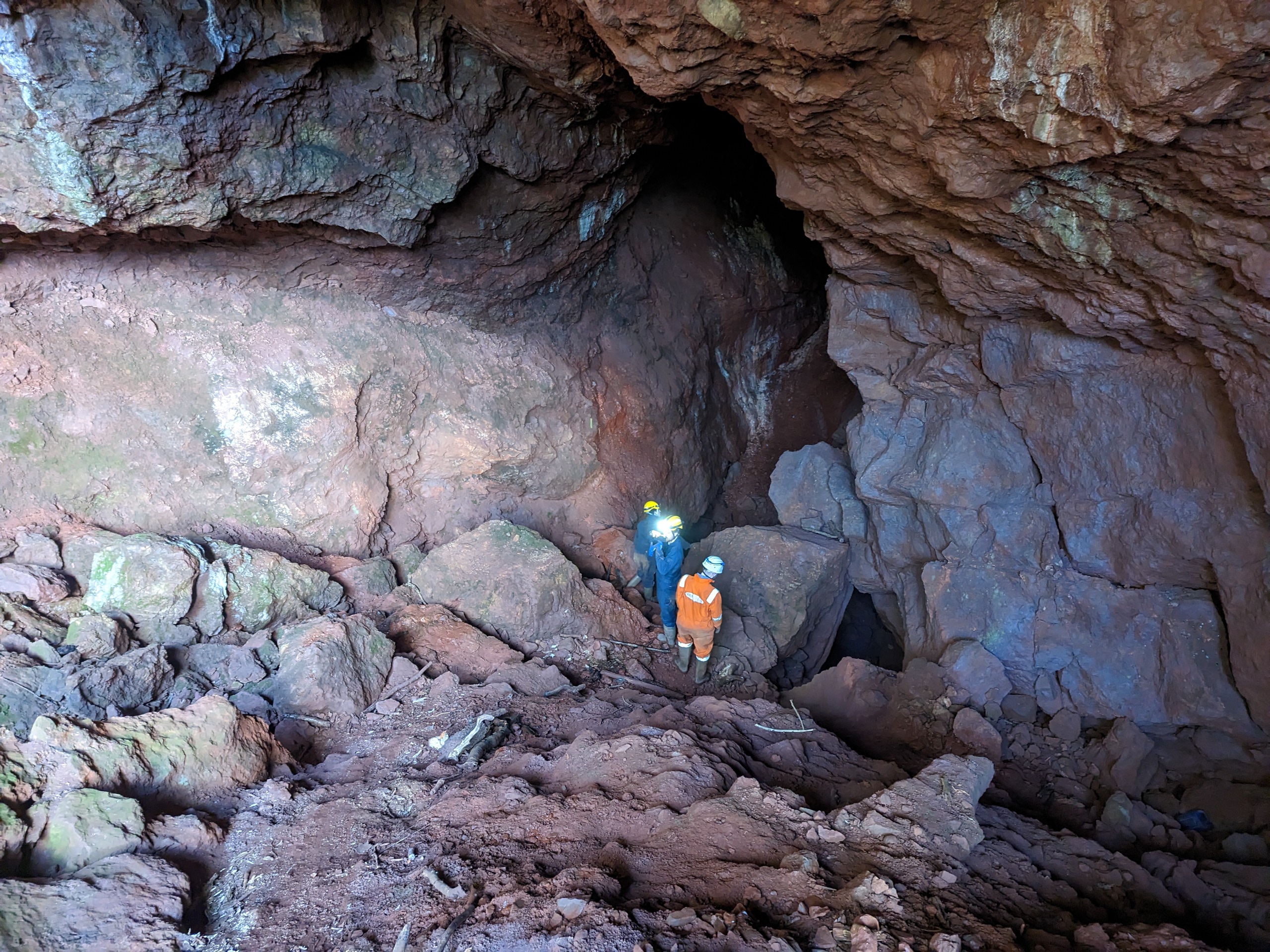 Two people in caving gear stood outside the front of a cave entrance.