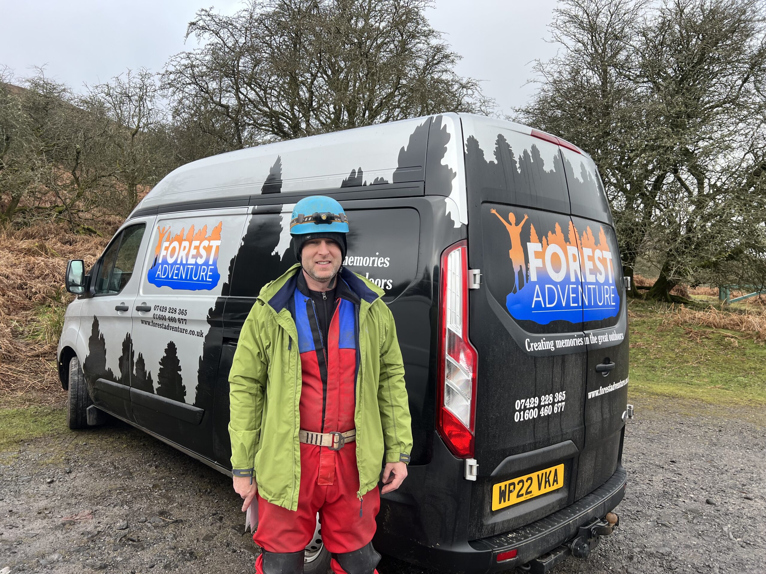 A man in red overalls and a blue helmet stood in front of a company van in a forest car park.