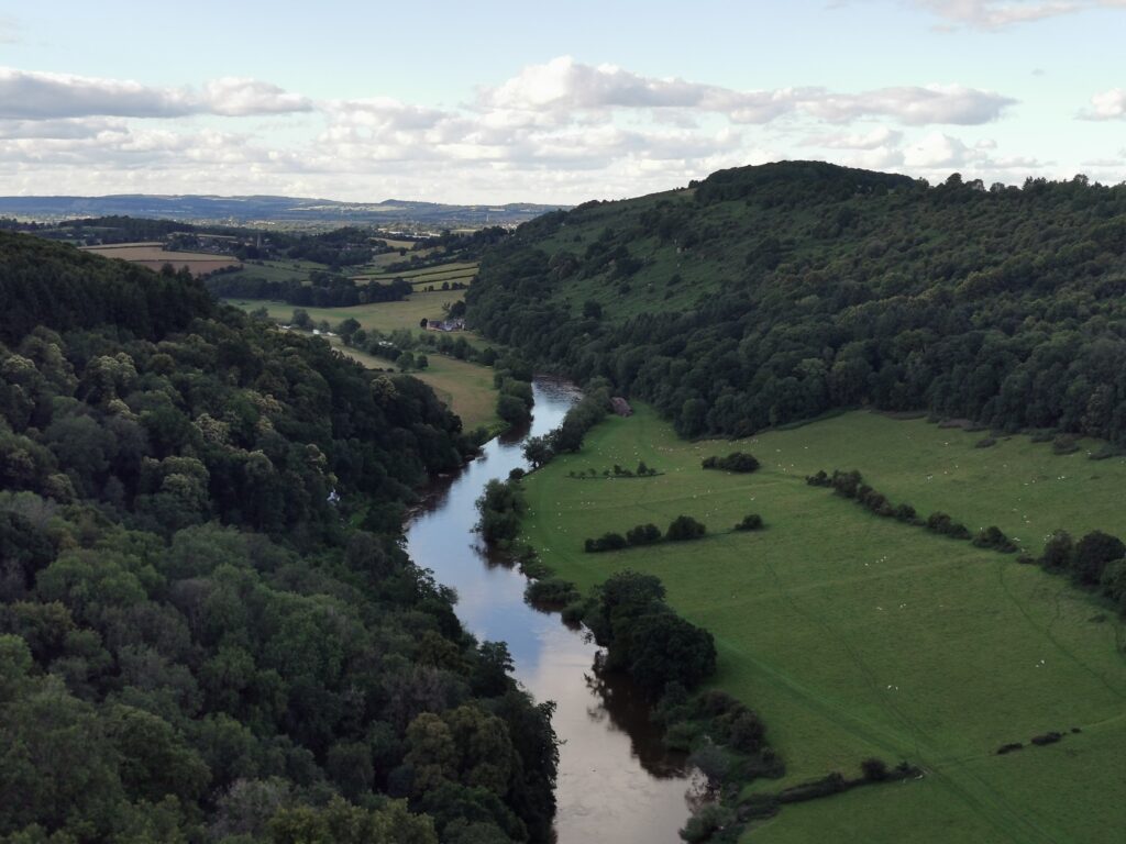A winding river flows through a lush green valley, bordered by wooded hills and open pastureland dotted with fields, under a partly cloudy sky.