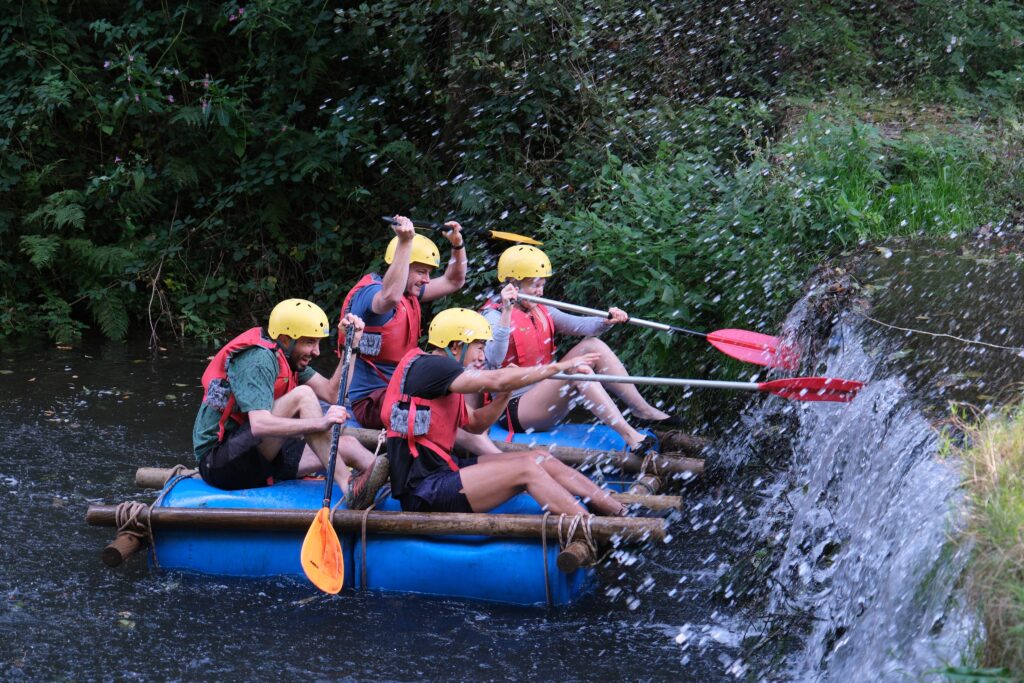 A group of four people on a raft in water made of large blue barrels and logs wearing hard hats and life jackets pushing off a small waterfall with their paddles.