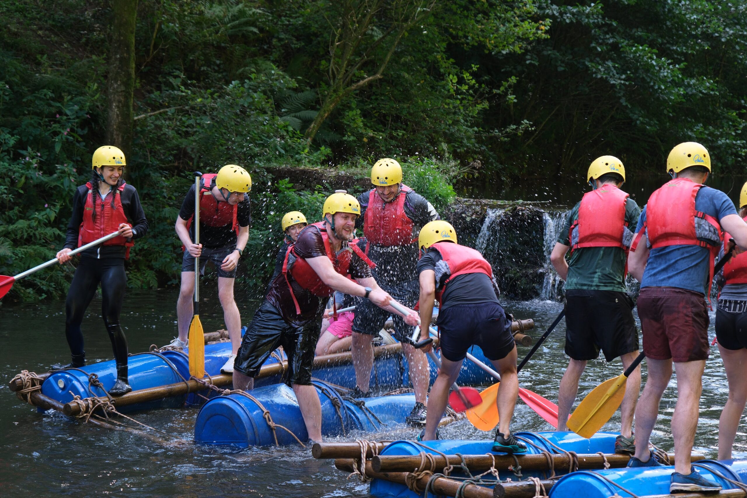 A group of people wearing helmets and buoyancy aids balance on barrel rafts in a shallow river, paddling and splashing water during a team-building rafting activity in a wooded setting.