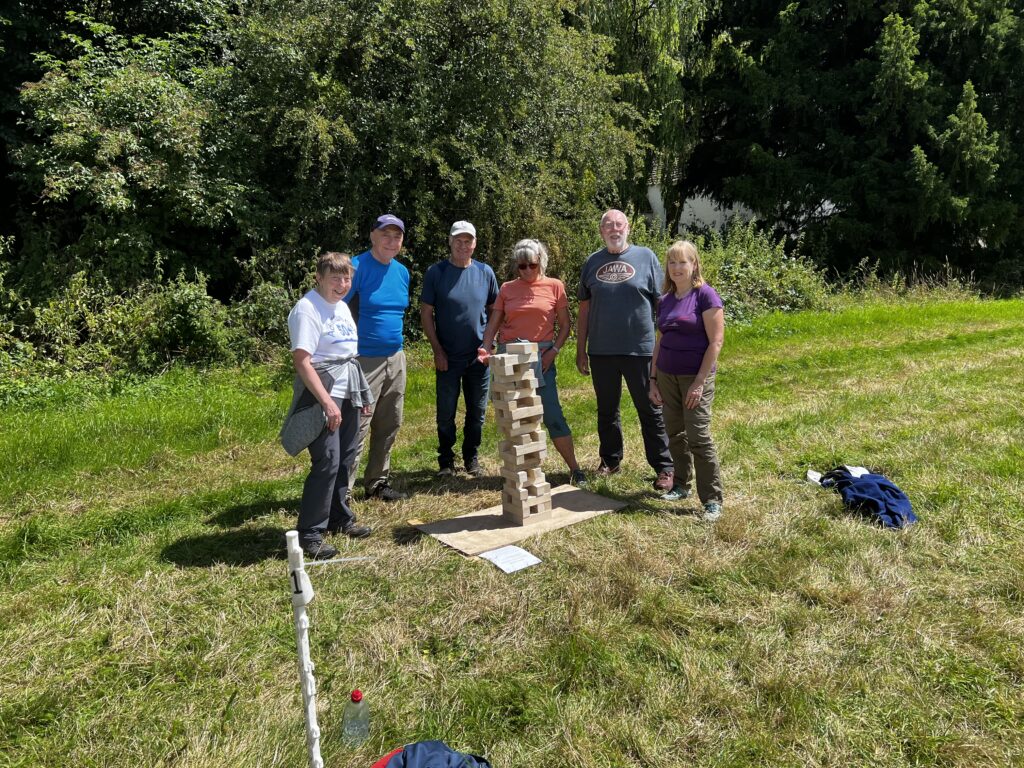 A group of adults gathered round a tall Jenga tower in a field.